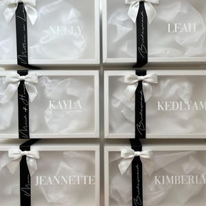 May include: White gift boxes with clear lids, each adorned with a white bow and a black ribbon. The boxes are labeled with names like "Nelly," "Leah," and "Kayla." The boxes are filled with white tissue paper.