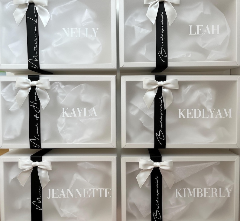 May include: White gift boxes with clear lids, each adorned with a white bow and a black ribbon. The boxes are labeled with names like "Nelly," "Leah," and "Kayla." The boxes are filled with white tissue paper.
