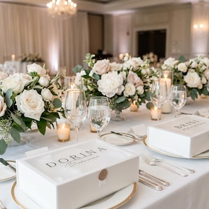 May include: A long, white table set for a formal event, adorned with floral centerpieces of white and blush roses, greenery, and lit candles. White boxes with gold accents and text are placed on the table, along with glassware and silverware.
