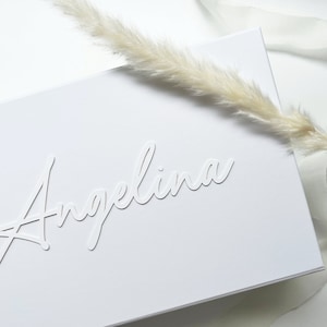 Peut inclure: Une carte blanche avec le nom "Angelina" en relief dans une police cursive. La carte est posée sur une surface blanche avec une plume blanche et un ruban blanc.