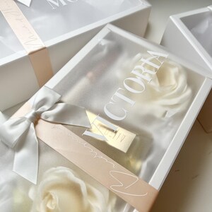 May include: White gift boxes with clear lids, tied with satin ribbons and bows. The boxes contain white roses and a gold-colored item. One box has the name "VICTORIA" printed on the lid.