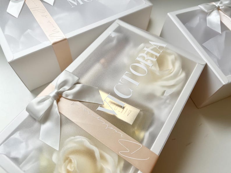 May include: White gift boxes with clear lids, tied with satin ribbons and bows. The boxes contain white roses and a gold-colored item. One box has the name "VICTORIA" printed on the lid.