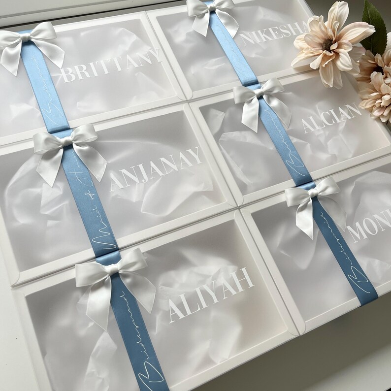 May include: White gift boxes with clear lids, each personalized with a name in white lettering. A light blue ribbon with white text and a white bow adorns each box. The boxes are arranged in a grid, suggesting a collection of gifts.