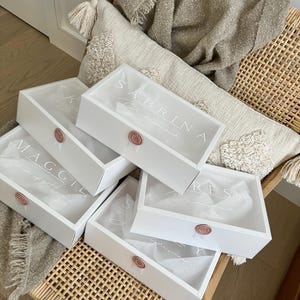 May include: White gift boxes with clear lids, each sealed with a rose-gold wax stamp. The boxes are personalized with names in elegant script, including "Sabrina" and "Maggie". The boxes are filled with tissue paper and are on a woven chair.