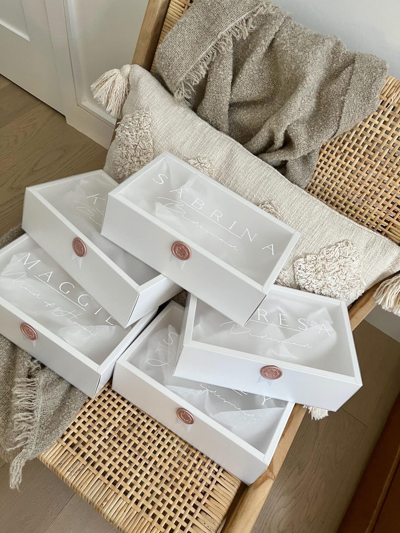 May include: White gift boxes with clear lids, each sealed with a rose-gold wax stamp. The boxes are personalized with names in elegant script, including "Sabrina" and "Maggie". The boxes are filled with tissue paper and are on a woven chair.