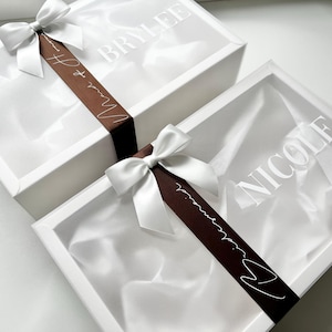 May include: Two white gift boxes with satin ribbon bows. One box has the name "BRYLEE" printed on the lid, and the other has "NICOLE". A brown ribbon with white text that reads "Maid of Honor" is wrapped around the boxes.