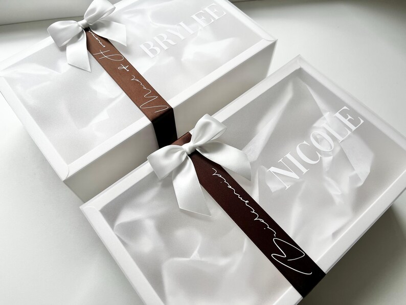 May include: Two white gift boxes with satin ribbon bows. One box has the name "BRYLEE" printed on the lid, and the other has "NICOLE". A brown ribbon with white text that reads "Maid of Honor" is wrapped around the boxes.
