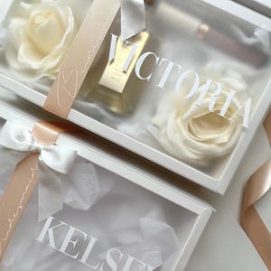 May include: White gift boxes with clear lids, each containing a cream-colored rose, a bottle of perfume, and a ribbon. The boxes are labeled with names in white script, and the ribbons say "bridesmaid".