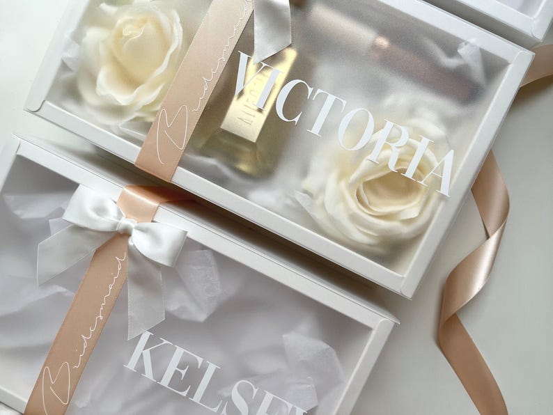 May include: White gift boxes with clear lids, each containing a cream-colored rose, a bottle of perfume, and a ribbon. The boxes are labeled with names in white script, and the ribbons say "bridesmaid".