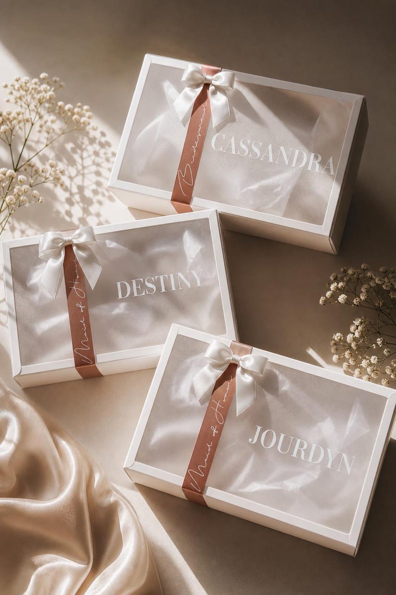 May include: White gift boxes with clear lids, each adorned with a white satin bow and a ribbon. The boxes are labeled with names: Cassandra, Destiny, and Jourdyn. The ribbon reads "Maid of Honor".