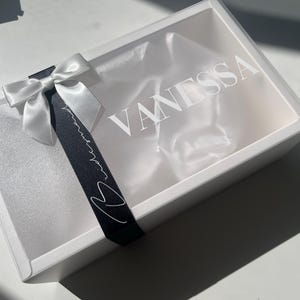 May include: A white gift box with a clear lid, featuring the name "VANESSA" in white lettering. A white satin bow and a black ribbon with white script and a heart accent the box. The box is empty.