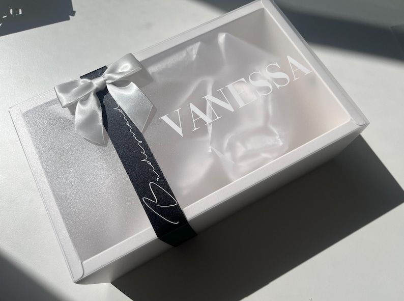 May include: A white gift box with a clear lid, featuring the name "VANESSA" in white lettering. A white satin bow and a black ribbon with white script and a heart accent the box. The box is empty.