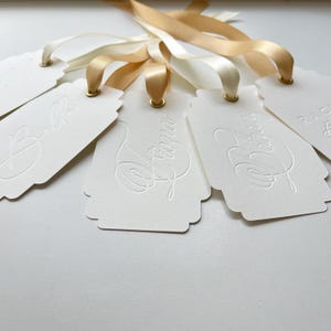 Peut inclure: Un ensemble d'étiquettes cadeaux blanches festonnées avec des noms en relief, notamment Bella, Fiona et Bethany. Chaque étiquette possède un œillet métallique doré et est fixée à un ruban de satin doré ou crème, formant un ensemble assorti.