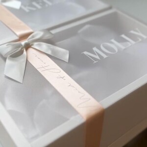 May include: Two white gift boxes with clear lids are tied with a silver ribbon and a peach-colored ribbon. The word "Molly" is printed on the lid of one box. The other box has the name "Keli" printed on the lid.