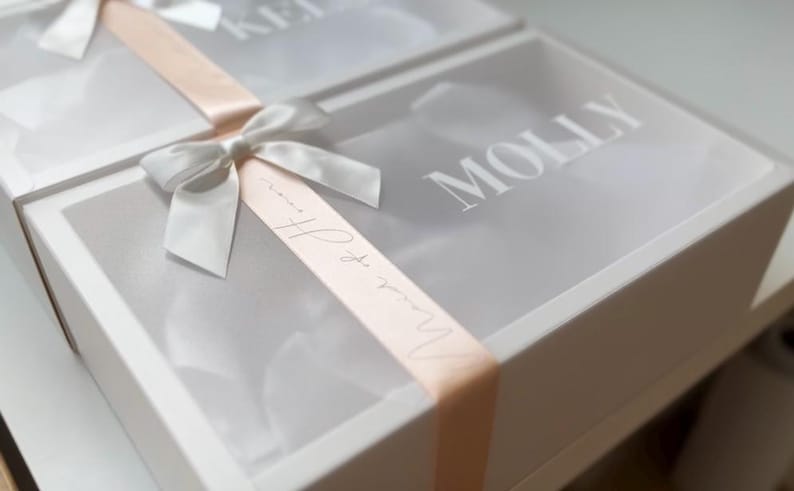 May include: Two white gift boxes with clear lids are tied with a silver ribbon and a peach-colored ribbon. The word "Molly" is printed on the lid of one box. The other box has the name "Keli" printed on the lid.