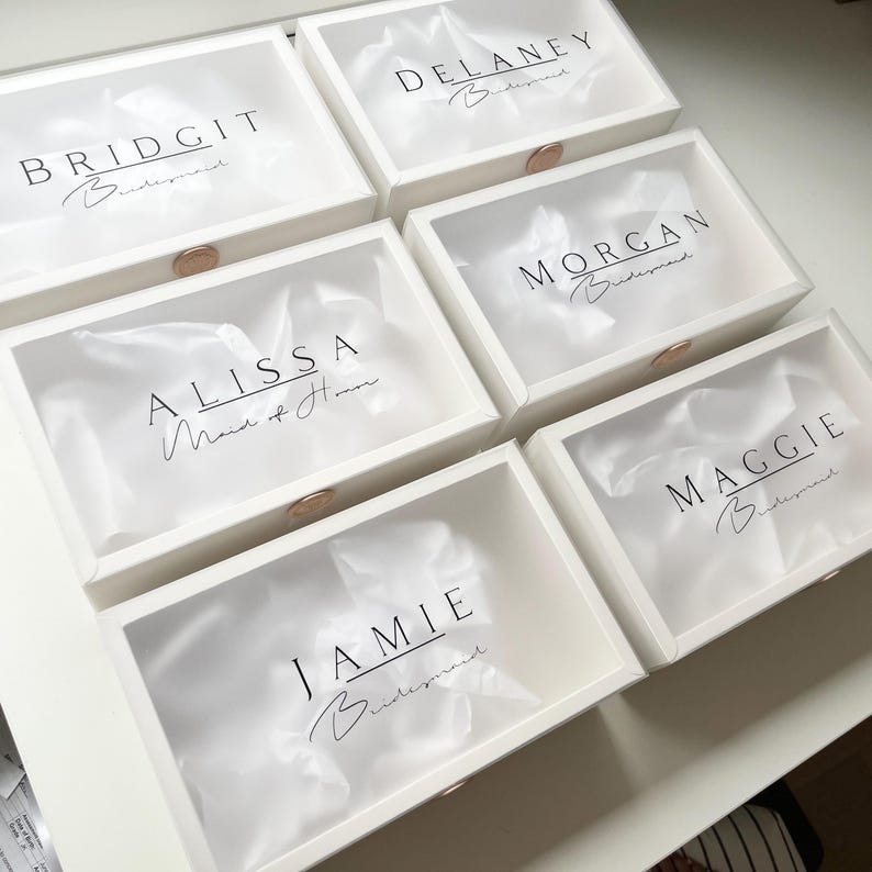May include: Six white gift boxes with clear lids, each labeled with a name in black script: Bridgit, Delaney, Alissa, Morgan, Maggie, and Jamie. The word "Bridesmaid" is written below each name. Each box has a gold-colored clasp.