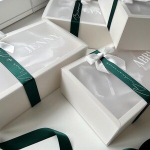 May include: White gift boxes with clear lids are tied with dark green satin ribbons and white bows. The boxes have names printed on the lids. The ribbons say "Maid of Honor" and "Bridesmaid".