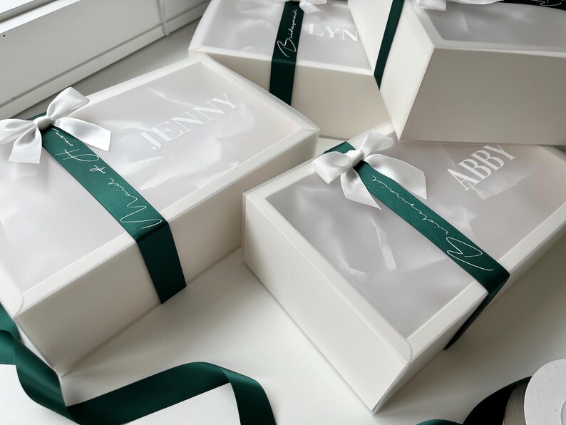 May include: White gift boxes with clear lids are tied with dark green satin ribbons and white bows. The boxes have names printed on the lids. The ribbons say "Maid of Honor" and "Bridesmaid".