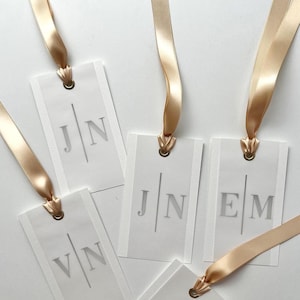 May include: White rectangular gift tags with gold satin ribbons. Each tag has a gold eyelet and features initials in a simple, elegant font. The initials are "JN", "VN", and "EM".