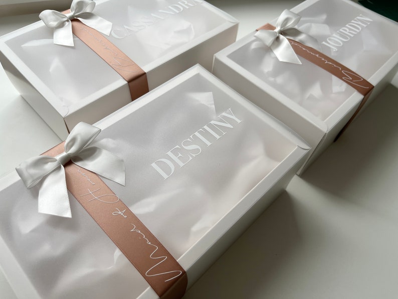 May include: Three white gift boxes with clear lids are arranged on a white surface. Each box is adorned with a white satin bow and a blush-colored ribbon. The names "Cassandre", "Jourdyn", and "Destiny" are printed on the boxes.