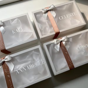 May include: Four white gift boxes with clear lids, each tied with a white satin bow and a copper-colored ribbon. The names "KALI", "CELESTE", "KRISTI", and "ANNABELLE" are printed on the boxes. The boxes are arranged on a white surface.