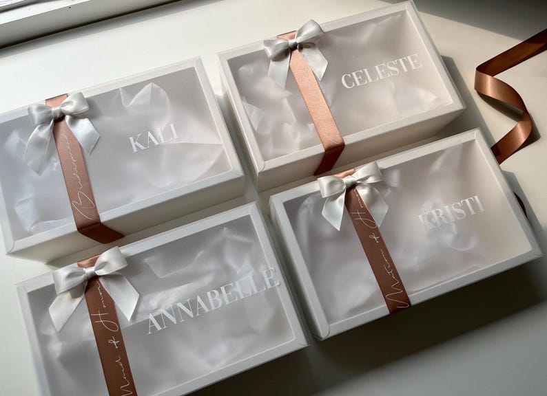 May include: Four white gift boxes with clear lids, each tied with a white satin bow and a copper-colored ribbon. The names "KALI", "CELESTE", "KRISTI", and "ANNABELLE" are printed on the boxes. The boxes are arranged on a white surface.