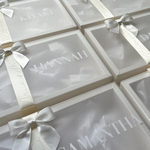 May include: Several white gift boxes with clear lids are arranged in a grid. Each box is adorned with a silver satin bow and a ribbon. The boxes have names printed on them, such as "Hannah" and "Samantha".