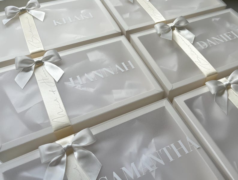 May include: Several white gift boxes with clear lids are arranged in a grid. Each box is adorned with a silver satin bow and a ribbon. The boxes have names printed on them, such as "Hannah" and "Samantha".
