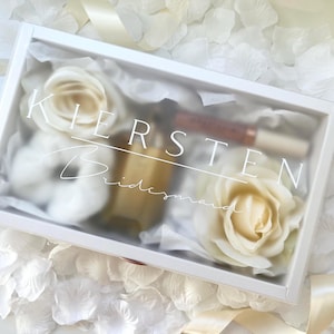 May include: A white gift box with the name "Kirsten" and the word "Bridesmaid" printed on the lid. Inside are white roses, a small bottle, and a tube of lip gloss. The box is surrounded by white flower petals.