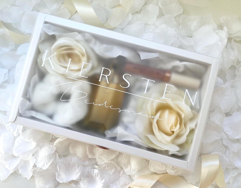 May include: A white gift box with the name "Kirsten" and the word "Bridesmaid" printed on the lid. Inside are white roses, a small bottle, and a tube of lip gloss. The box is surrounded by white flower petals.
