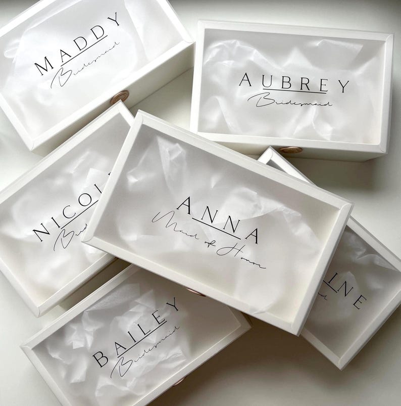 May include: White gift boxes with clear lids, each labeled with a name in black script. Some boxes include the word "Bridesmaid" or "Maid of Honor". The boxes are filled with white tissue paper.