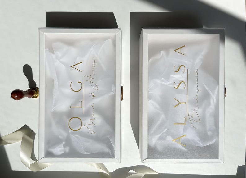 May include: Two white rectangular boxes with gold lettering and a brown handle. The box on the left says "OLGA Maid of Honor" and the box on the right says "ALYSSA Bridesmaid". Both boxes are filled with white tissue paper.