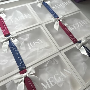 May include: White gift boxes with clear lids, each adorned with a satin bow and ribbon. The ribbons are either burgundy or navy blue, with white script that reads "Maid of Honor" and names like "Tasha" and "Josy" on the box lids.