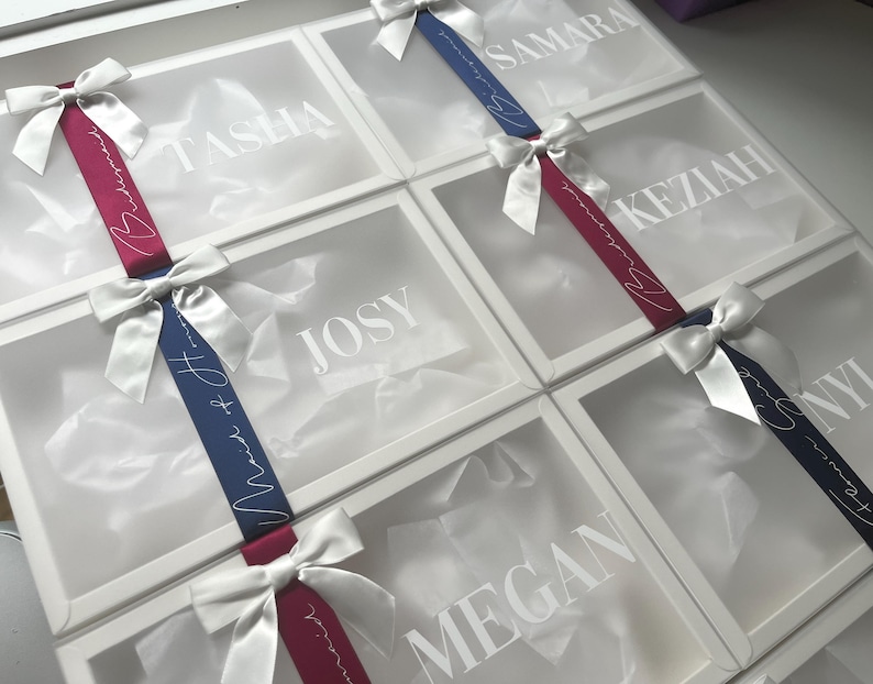 May include: White gift boxes with clear lids, each adorned with a satin bow and ribbon. The ribbons are either burgundy or navy blue, with white script that reads "Maid of Honor" and names like "Tasha" and "Josy" on the box lids.