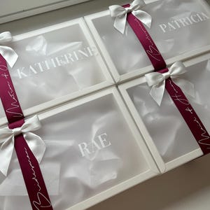 May include: Four white gift boxes with clear lids, each adorned with a white satin bow and a burgundy ribbon. The boxes are labeled with names in white lettering: Katherine, Patricia, Jane, and Rae. The ribbon has white script.