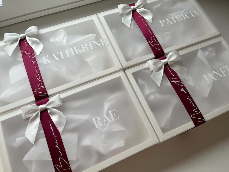 May include: Four white gift boxes with clear lids, each adorned with a white satin bow and a burgundy ribbon. The boxes are labeled with names in white lettering: Katherine, Patricia, Jane, and Rae. The ribbon has white script.