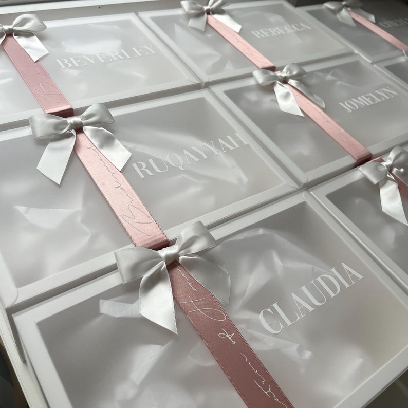 May include: White gift boxes with clear lids, each adorned with a white bow and a pink ribbon. The lids are printed with names in a white font. The boxes are arranged in a grid pattern.