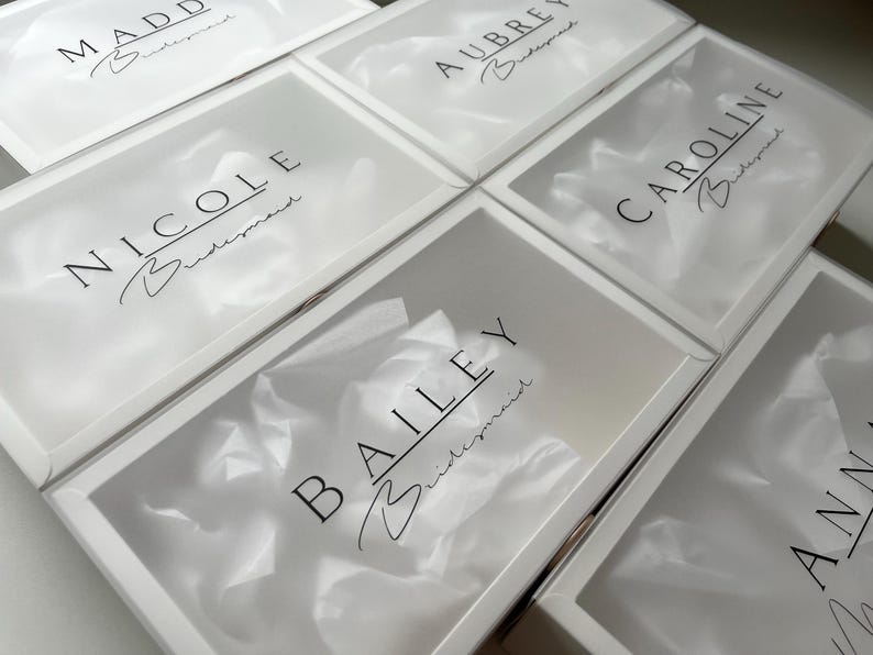May include: Several white gift boxes with clear lids are arranged in a grid. Each box has a name printed in black script, with the word "Bridesmaid" below. The boxes are filled with white tissue paper.