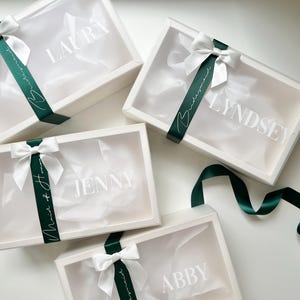 May include: Four white gift boxes with clear lids, each adorned with a white bow and a dark green ribbon. The boxes are labeled with names in white lettering: Laura, Lyndsey, Jenny, and Abby. The ribbon says "Bridesmaid" and "Maid of Honor".