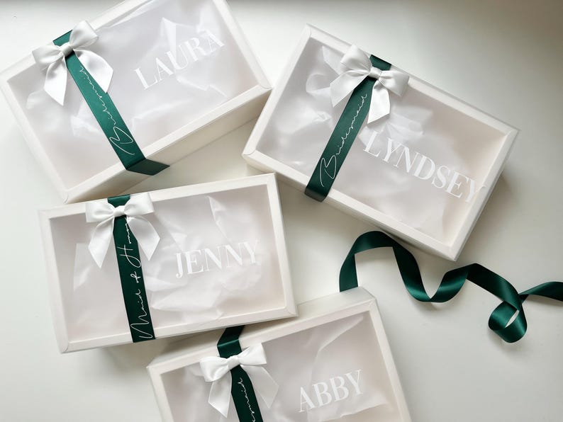 May include: Four white gift boxes with clear lids, each adorned with a white bow and a dark green ribbon. The boxes are labeled with names in white lettering: Laura, Lyndsey, Jenny, and Abby. The ribbon says "Bridesmaid" and "Maid of Honor".