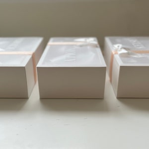 May include: Three white rectangular boxes with lids, arranged in a row. Each box is wrapped with a light pink ribbon and a white bow. The lids have text printed on them. The boxes are on a white surface.