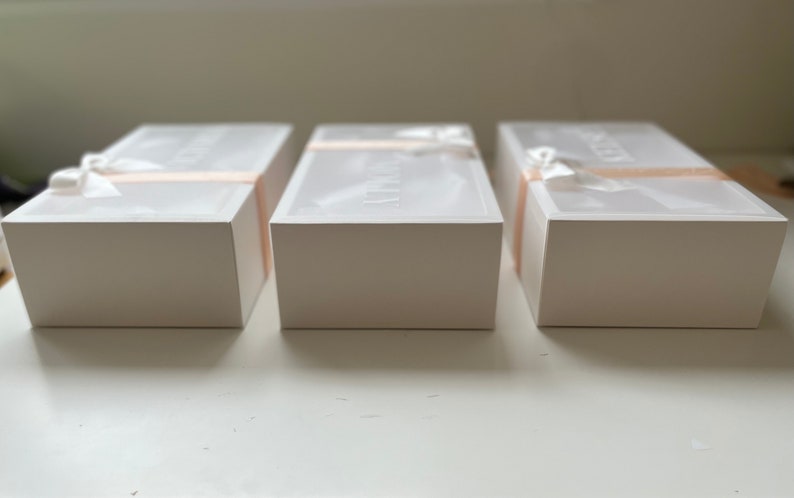 May include: Three white rectangular boxes with lids, arranged in a row. Each box is wrapped with a light pink ribbon and a white bow. The lids have text printed on them. The boxes are on a white surface.