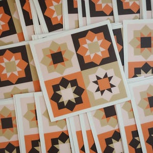 May include: A stack of square greeting cards featuring a geometric star pattern in shades of orange, dark brown, and beige. Each card displays a four-quadrant design with alternating star motifs, printed on white cardstock with a clean border.