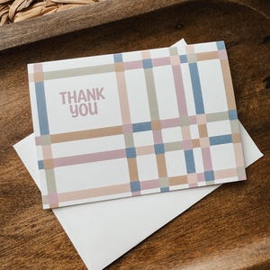 May include: A rectangular white greeting card featuring a grid pattern of intersecting horizontal and vertical lines in muted shades of blue, tan, and dusty pink. The text THANK YOU is printed in a soft pink sans-serif font on the left side.