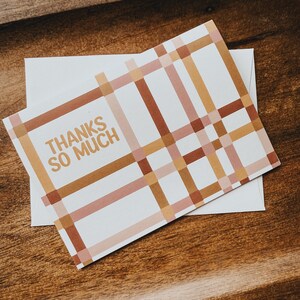 May include: A rectangular white greeting card featuring a grid pattern of intersecting horizontal and vertical lines in shades of brown, tan, and muted pink. The text THANKS SO MUCH is printed in a bold, sans-serif font on the left side.