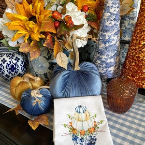 May include: A blue velvet pumpkin, a gold velvet pumpkin, and a white tea towel with a blue and white porcelain vase with a stack of white pumpkins and fall leaves. The pumpkins are on a blue and white checkered tablecloth.