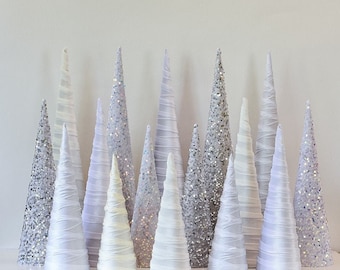 May include: A collection of white and silver cone-shaped Christmas trees with glitter and satin fabric. The trees are arranged in a row on a white surface.