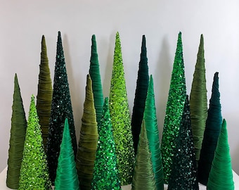 May include: A collection of green cone-shaped Christmas trees made of fabric and sequins. The trees are arranged in a row on a white surface.
