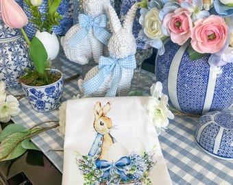 May include: A white tea towel featuring a blue and white illustration of a bunny rabbit holding an egg in a blue and white floral basket. The towel is laid on a blue and white gingham tablecloth.