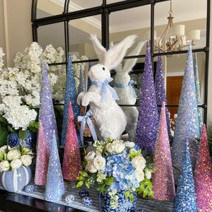 May include: A white faux fur bunny rabbit figurine with a blue and white ribbon sits in front of a mirror with a black frame.  The bunny is surrounded by sparkly cone-shaped decorations in shades of blue, pink, and purple.  A blue and white floral arrangement is in a blue and white ceramic vase.